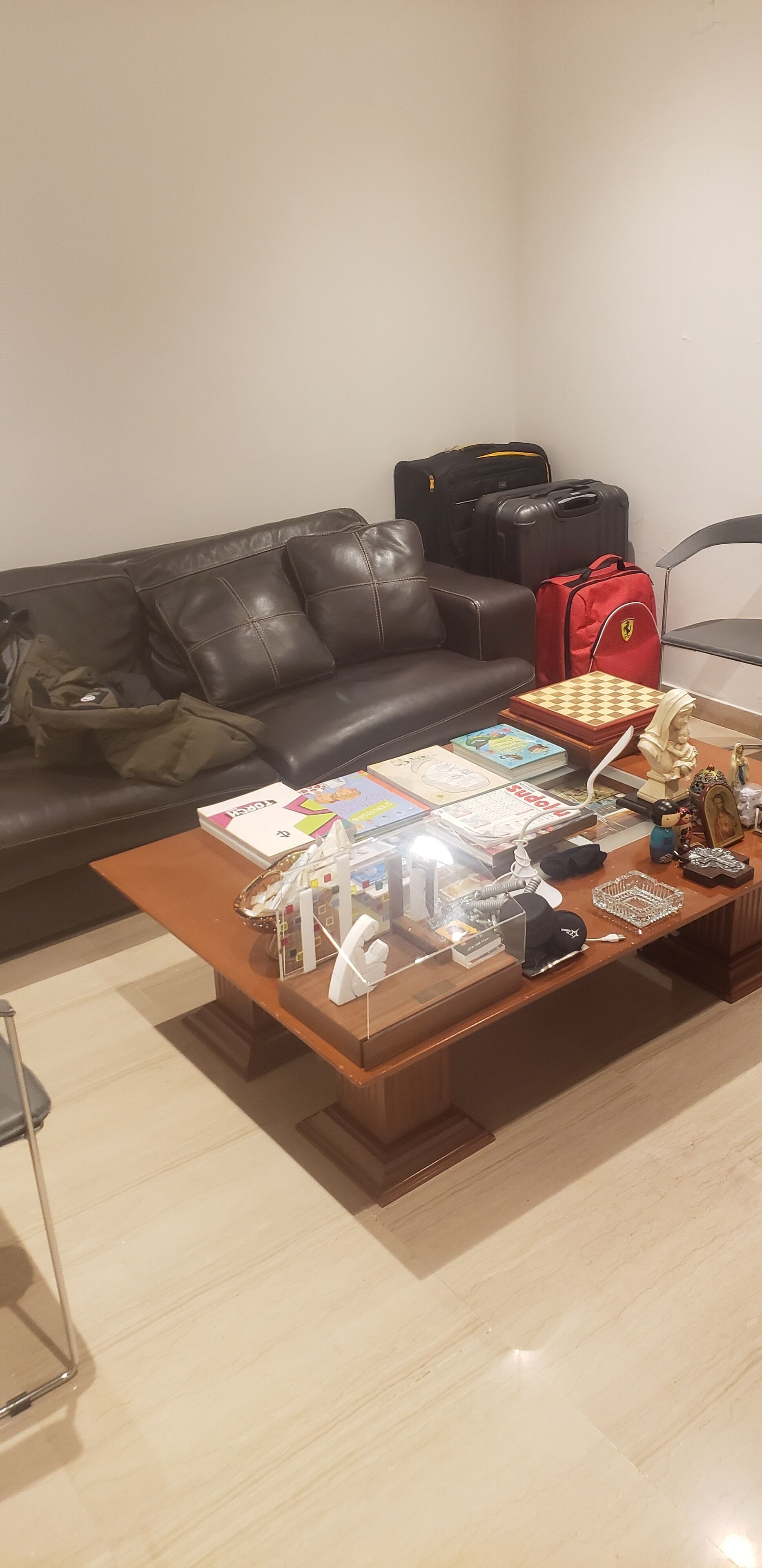 A living room with a brown leather sofa, a wooden coffee table with various items, and luggage in the background.