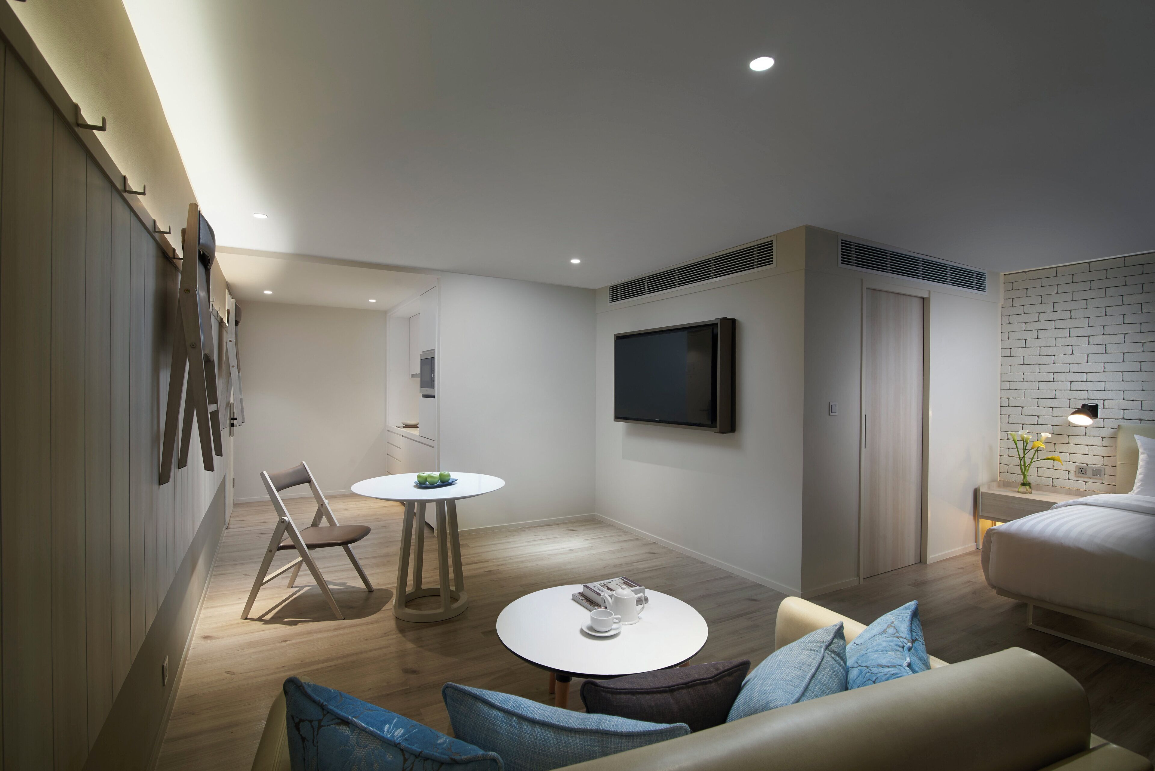 A modern hotel room with a bed, a TV mounted on the wall, a small round table with a chair, and a sofa with cushions.