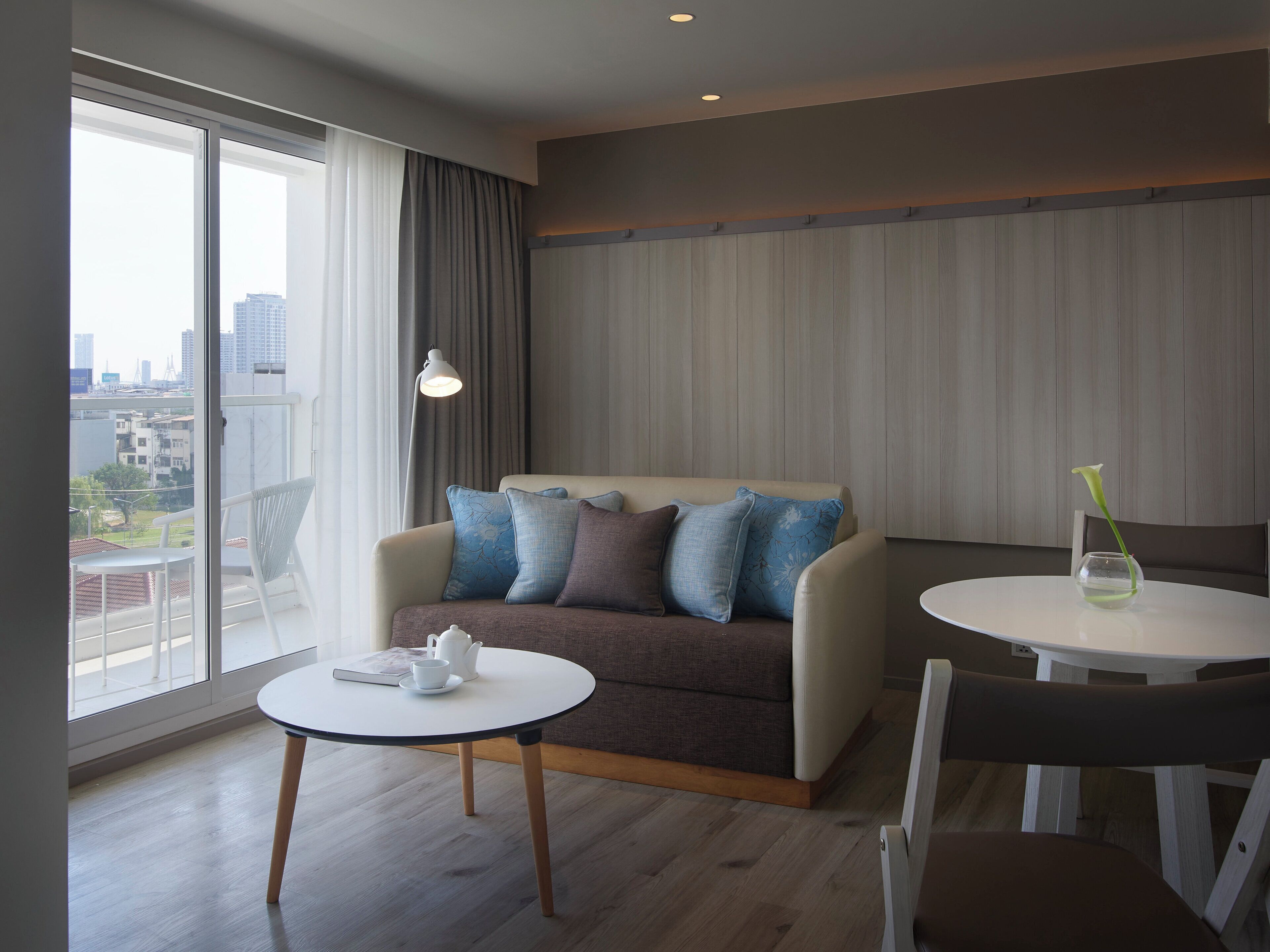 A modern living room with a sofa, a round table, and a chair. There is a view of the city through large windows.