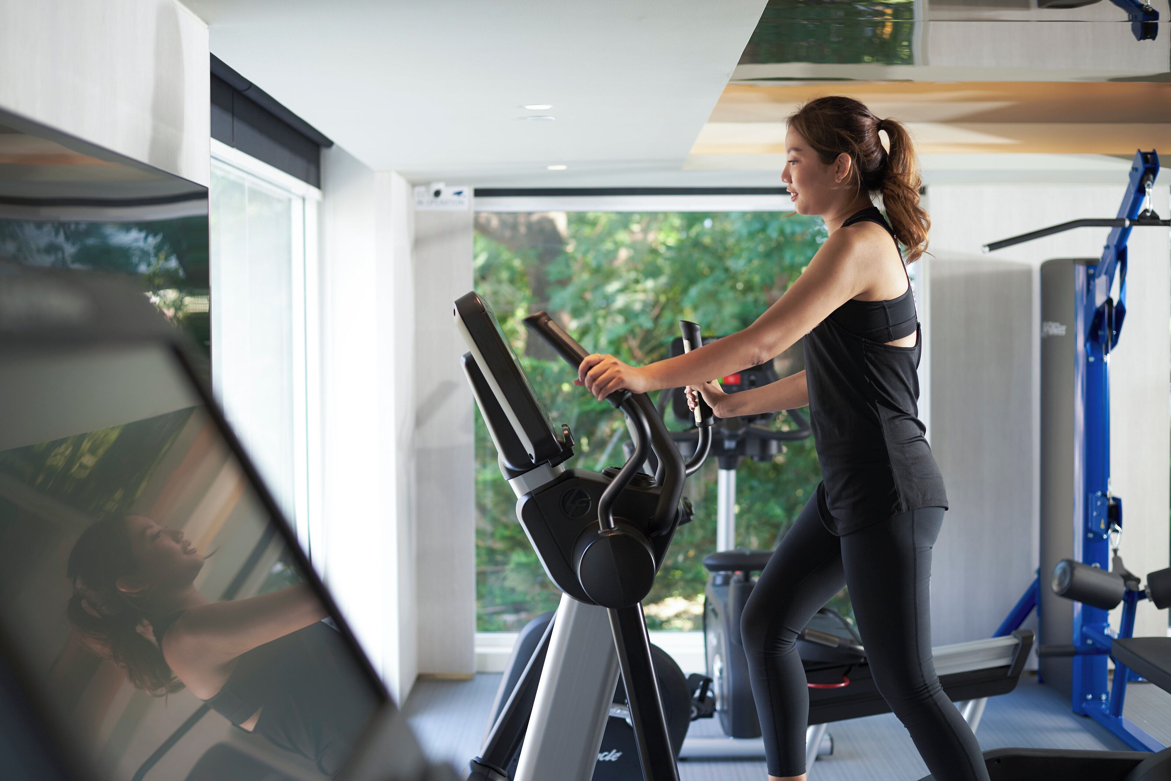 A woman using an elliptical trainer in a gym with large windows and exercise equipment.