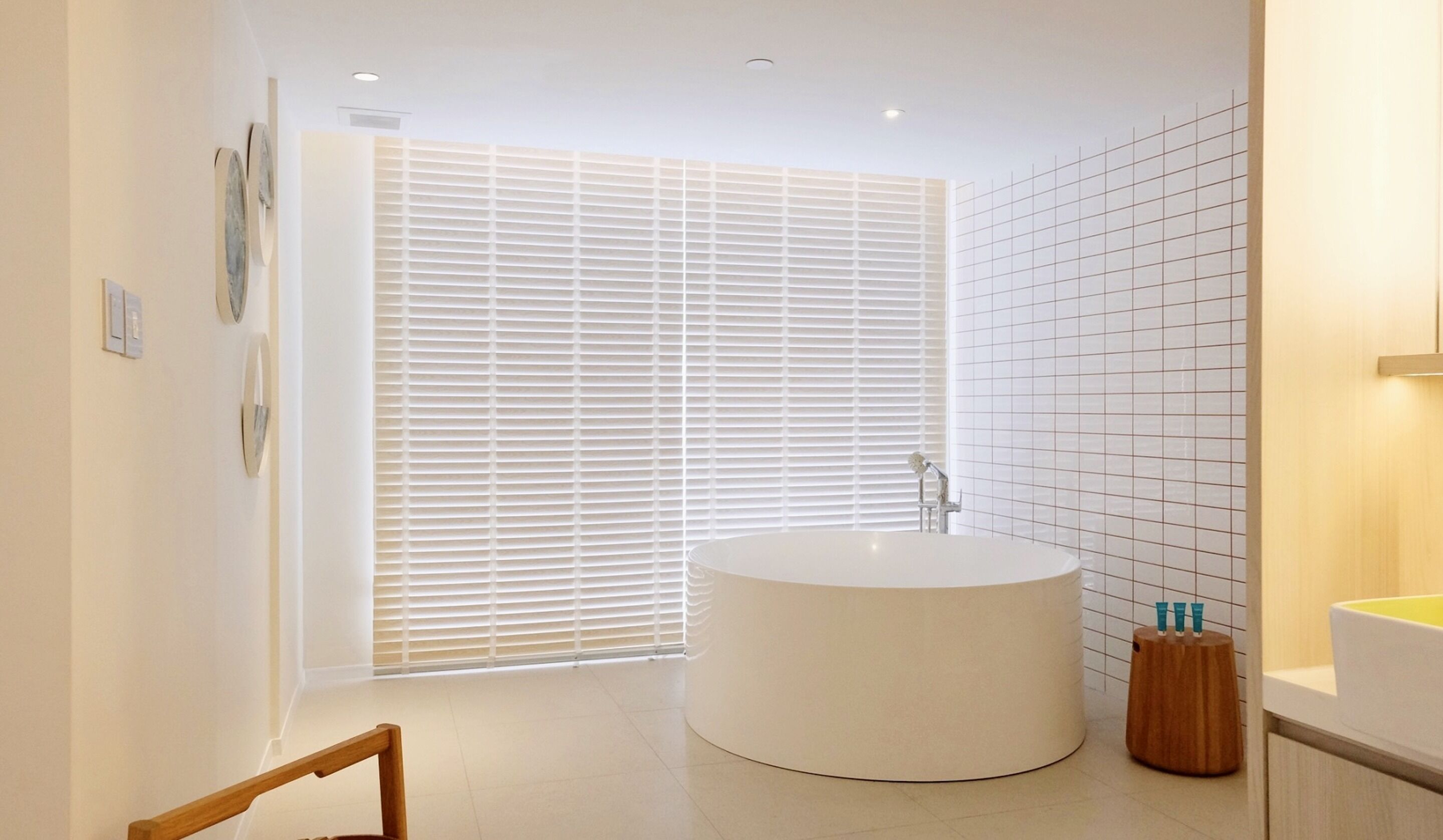 A modern bathroom with a freestanding bathtub, a large window with blinds, and a tiled wall.