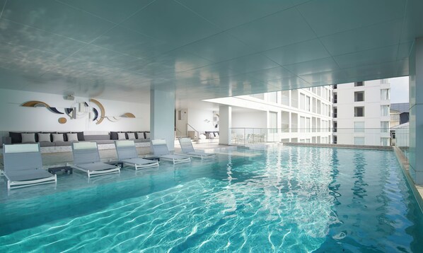 Een binnenzwembad met ligbedden, een modern plafondontwerp en een wit gebouw op de achtergrond.