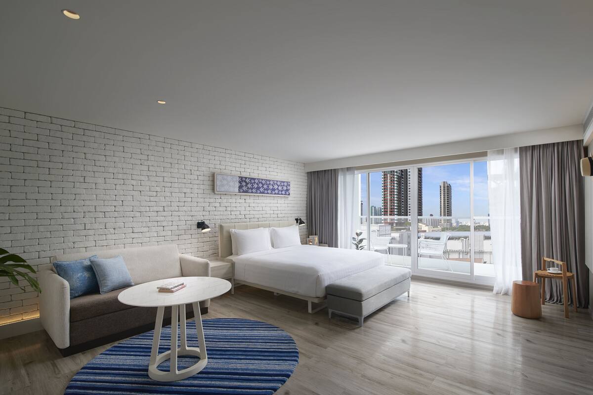 A modern hotel room with a large bed, a sofa, a small table, and a view of the city through large windows.
