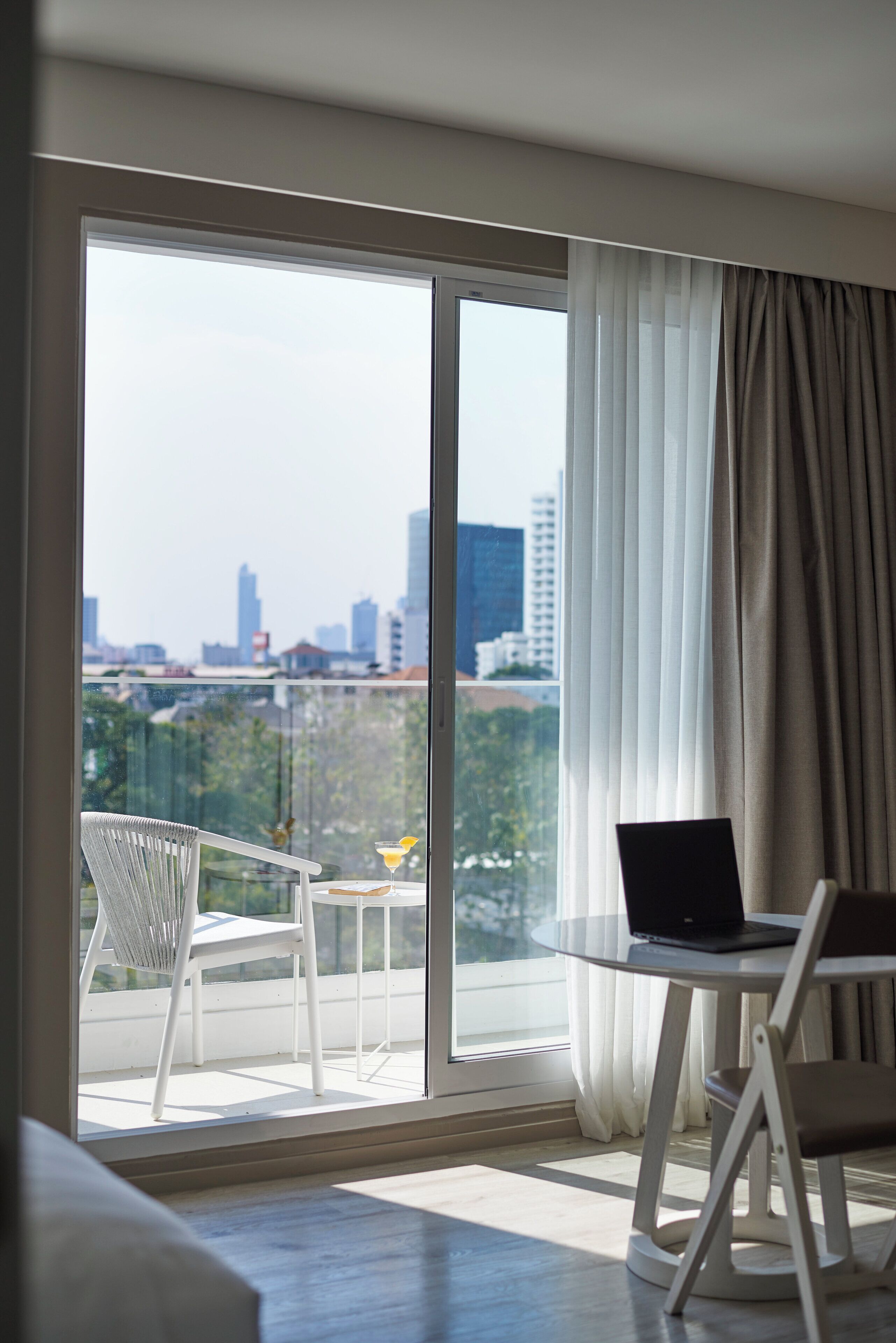 A modern room with a large sliding glass door leading to a balcony. A laptop is placed on a small round table, and there is a white chair on the balcony.