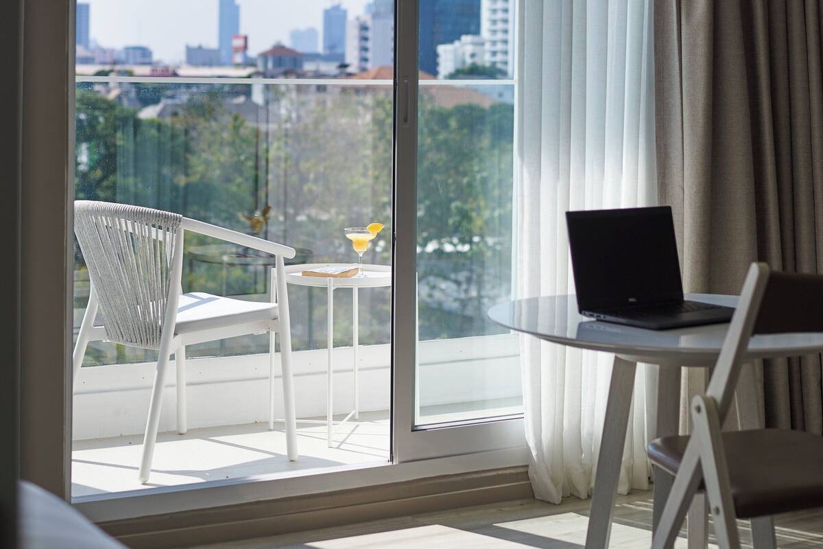 A modern room with a large sliding glass door leading to a balcony. A laptop is placed on a small round table, and there is a white chair on the balcony.