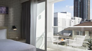 Een moderne hotelkamer met een groot bed, een balkon met witte stoelen en uitzicht op de stad.