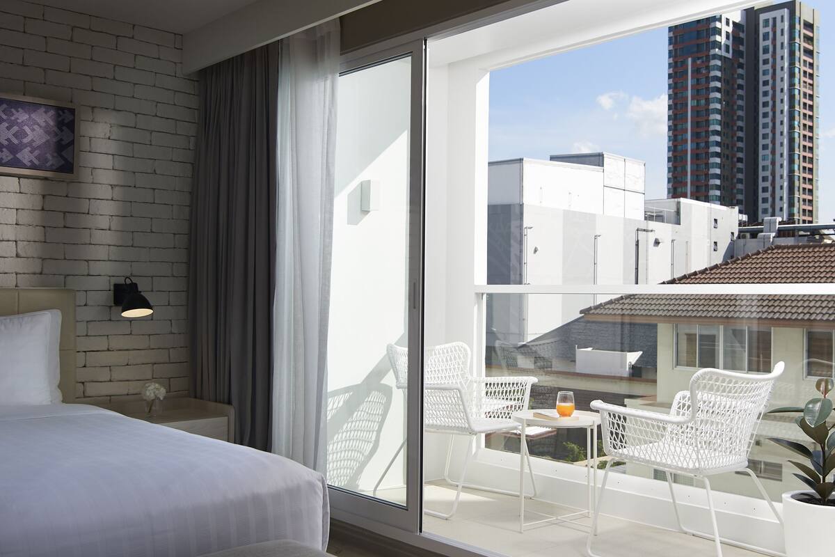 A modern hotel room with a large bed, a balcony with white chairs, and a city view.
