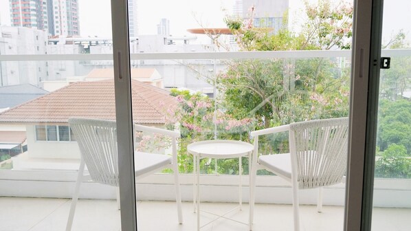 A balcony with two white chairs and a small table, overlooking a garden with pink flowers and a building in the background.
