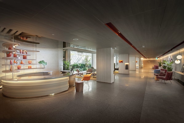 Un hall d’hôtel moderne doté d’un comptoir d’accueil central, d’un espace salon et de grandes fenêtres laissant entrer la lumière naturelle.