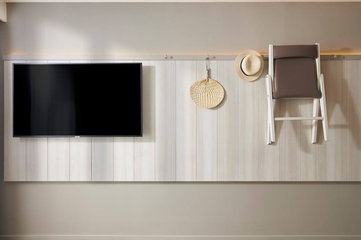 A modern living room with a flat-screen TV mounted on a light wood-paneled wall, a wicker wall hanging, and a brown leather chair.