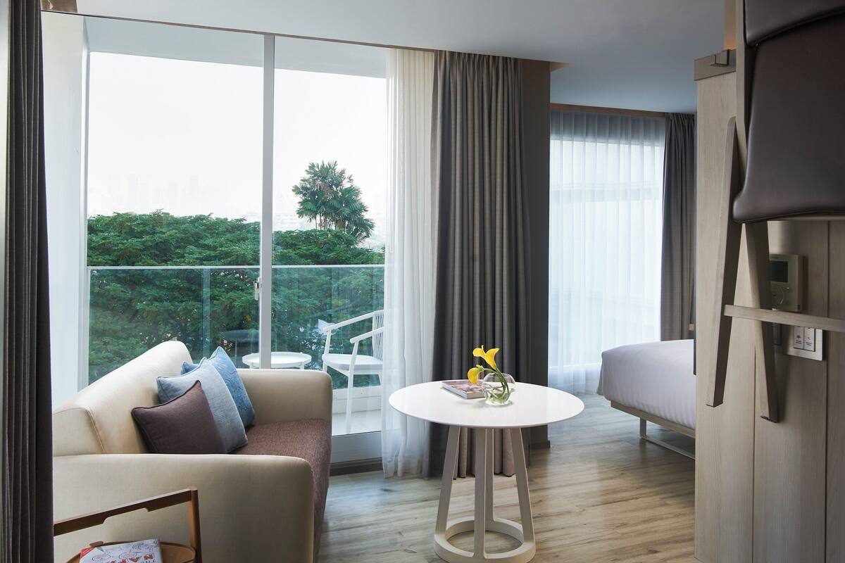 A modern hotel room with a sofa, a small table with a vase, a chair, and a bed.