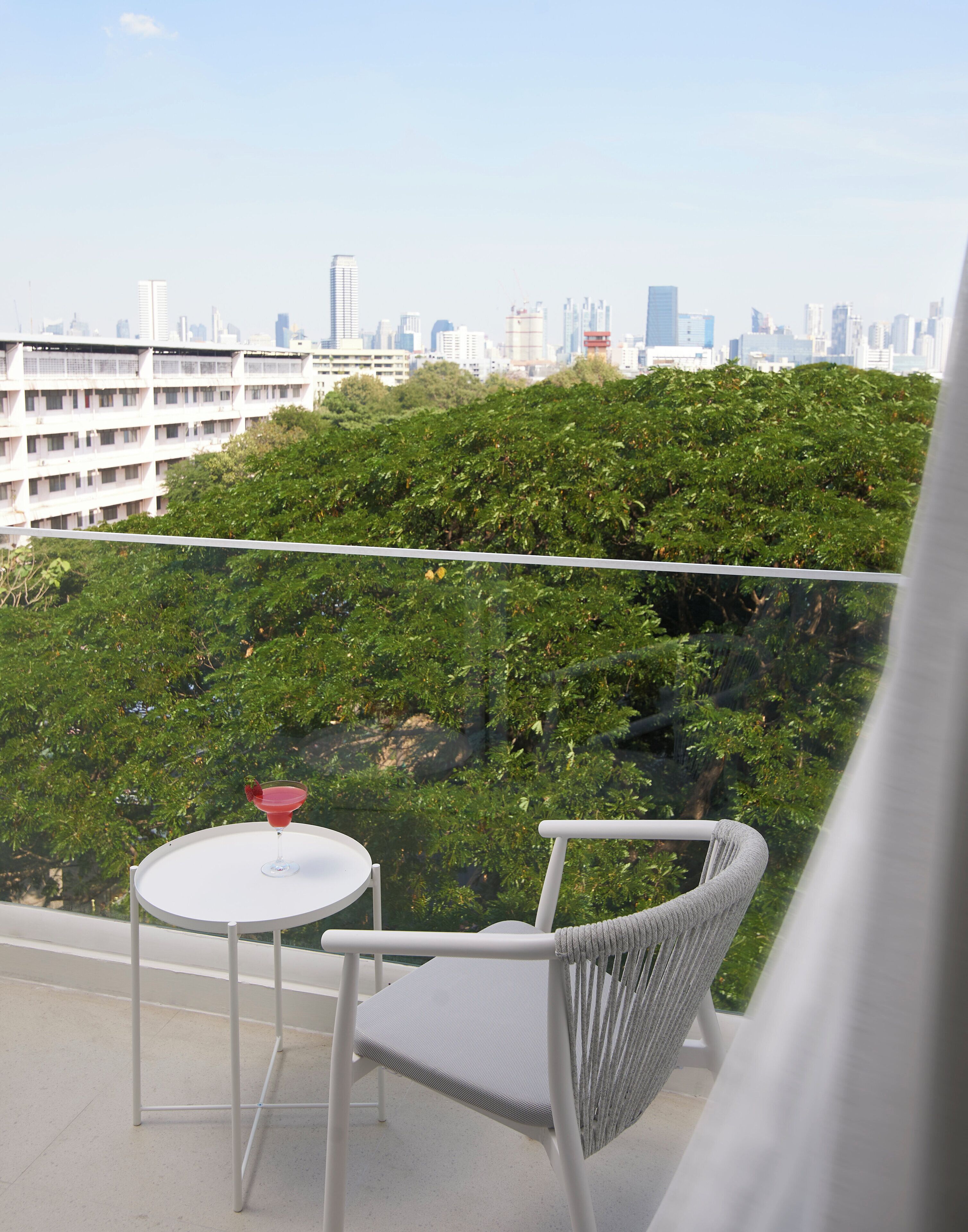 A balcony with a white table and chair, a drink on the table, and a cityscape view in the background.