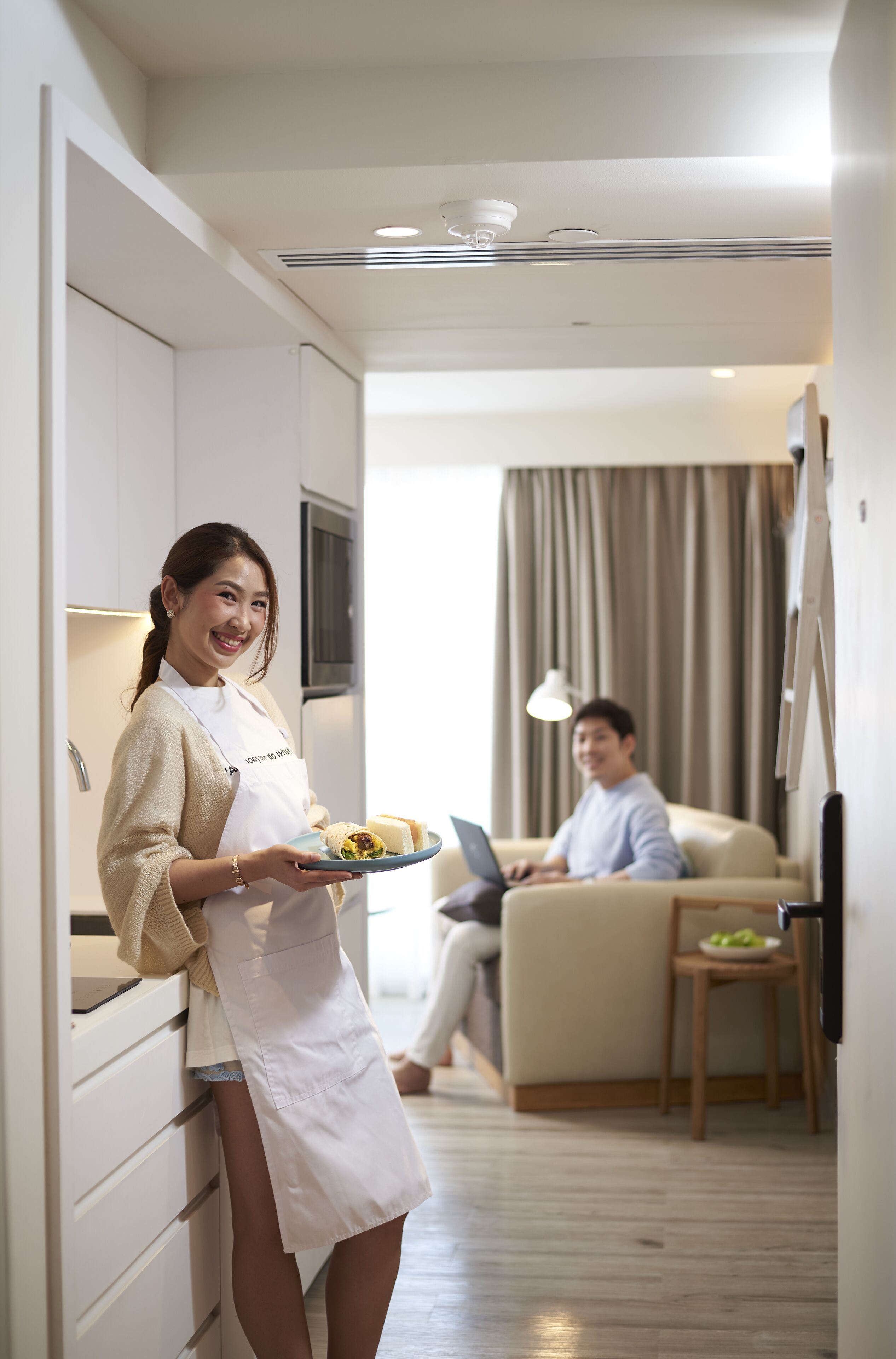 Une cuisine moderne avec des meubles blancs, une personne tenant une assiette de nourriture et, au fond, un homme utilisant un ordinateur portable.