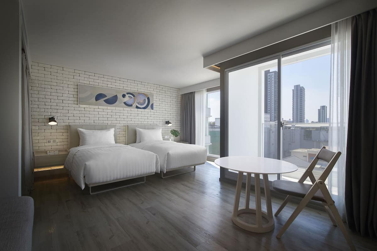A modern hotel room with two beds, a dining table, and a city view.