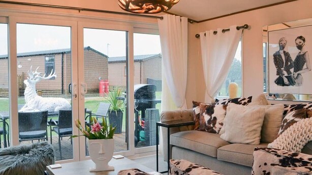 A modern living room with a sofa, a coffee table, and a fireplace. There is a large window with a view of a house and a deer statue outside. - Boutique Lodge With hot tub Sleeps 6 (Bridlington)