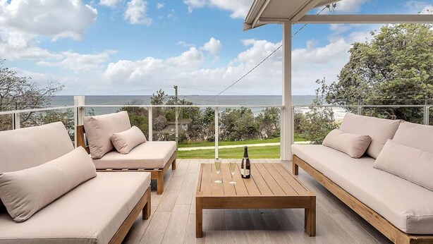 A balcony with a wooden table and two beige sofas, overlooking a coastal landscape. - Getawayz NSW -  'Drift' - Absolute Beachfront at Coledale w/ Panoramic Views & Multiple Living Zones (Coledale)