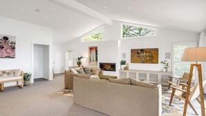 A spacious living room with a large beige sofa, a wooden coffee table, and a floor lamp. There are two windows providing natural light, and a painting is visible on the wall. - Getawayz NSW -  'Drift' - Absolute Beachfront at Coledale w/ Panoramic Views & Multiple Living Zones (Coledale)