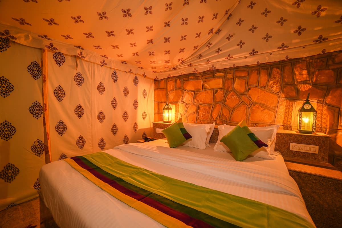 Una cama con una colcha de colores de arcoíris, almohadas blancas y almohadas decorativas verdes y púrpuras. La habitación tiene una pared de piedra y una cortina estampada. Hay dos lámparas fijadas a la pared a cada lado de la cama.