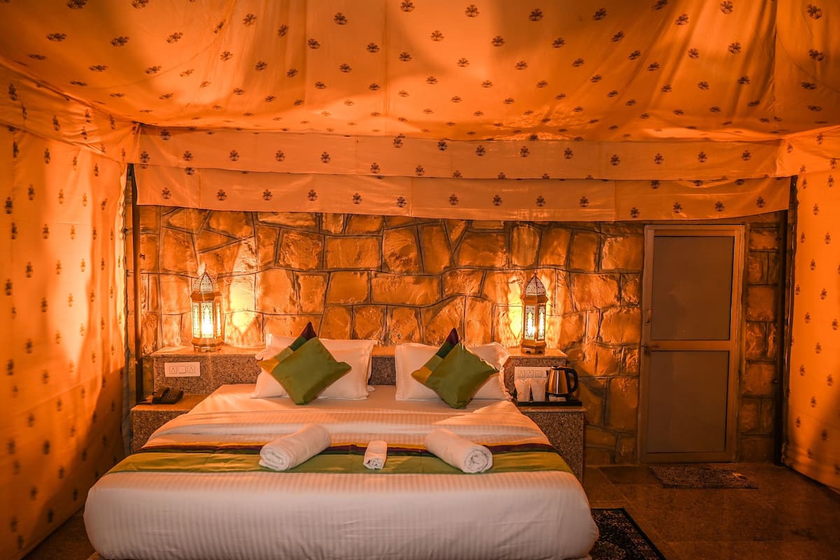 Una habitación con pared de piedra, una cama con almohadas, una mesita de noche, un farol y una puerta.