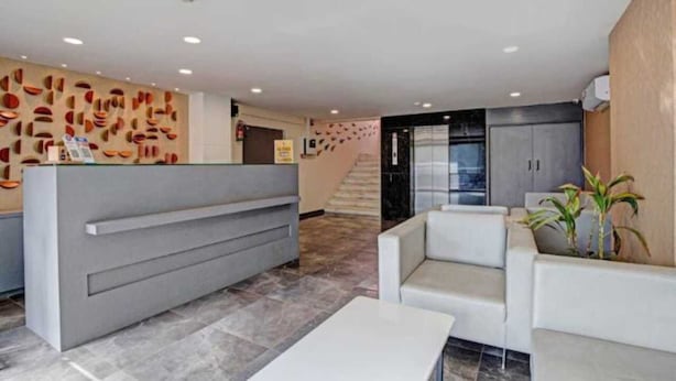 Un área de recepción moderna con un mostrador gris, zona de estar con sofás blancos y una pared decorativa con patrones abstractos.