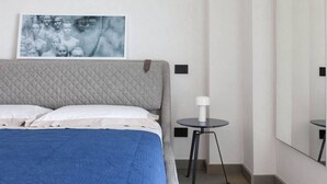 Een netjes opgemaakt bed met een gewatteerd hoofdbord, een nachtkastje met een lamp en een ingelijst beeld aan de muur.