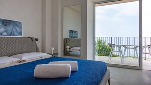 Een moderne slaapkamer met een groot bed, een balkon met witte stoelen en uitzicht op zee.