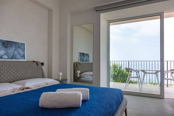 A modern bedroom with a large bed, a balcony with white chairs, and a view of the sea.