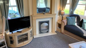 Una sala de estar con una televisión de pantalla plana sobre un mueble de madera, una chimenea, un sofá con cojines y una ventana con cortinas.
