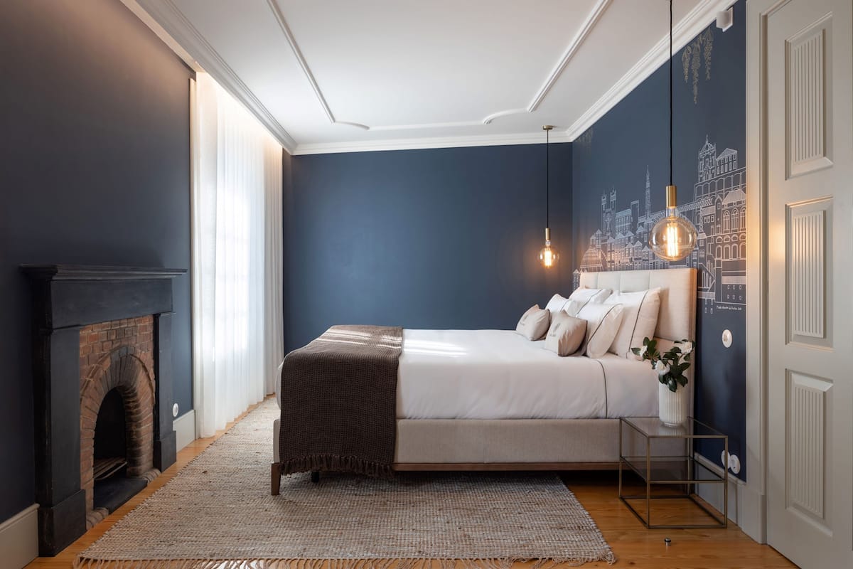 Une chambre avec un lit, une cheminée, un mur orné d’une fresque représentant un paysage urbain et un luminaire suspendu.