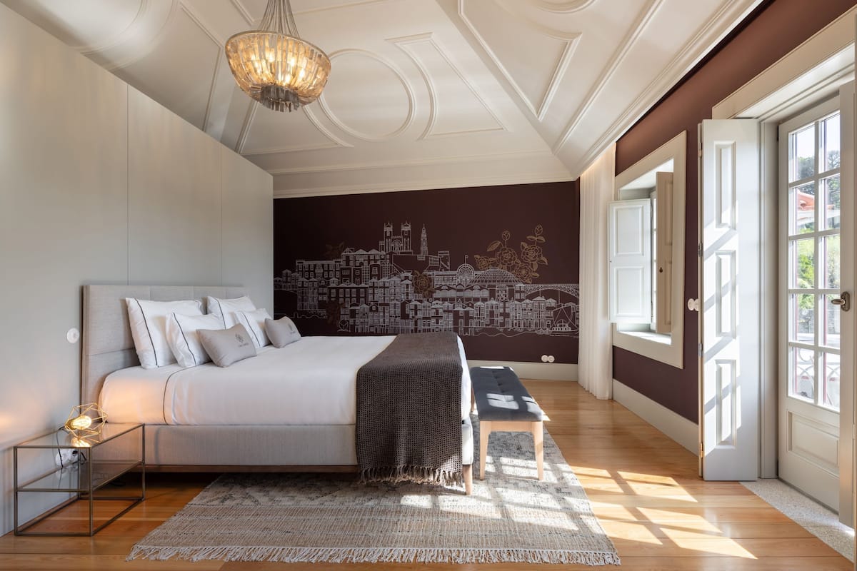 Une chambre avec une fresque représentant un paysage urbain, un lit avec des oreillers, une table de chevet avec une lampe et une grande fenêtre laissant entrer la lumière naturelle.