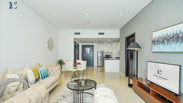 Un salon moderne avec un canapé, une table basse et un téléviseur. On aperçoit une cuisine au fond.