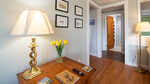A room with a wooden desk, a lamp, a vase with yellow flowers, framed pictures on the wall, and a door leading to another room.
