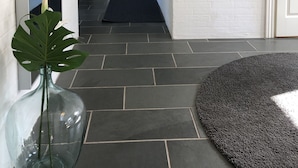 Un couloir recouvert d’une moquette noire mène à une pièce où se trouvent un lit et un bureau. Il y a une plante sur la gauche et un tableau accroché au mur.