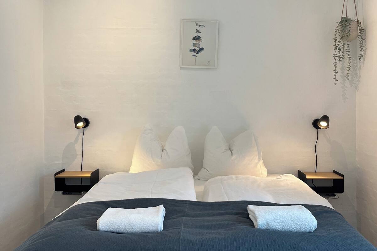 A bedroom with a bed, white pillows, a bedside table, a wall-mounted light, and a hanging plant.