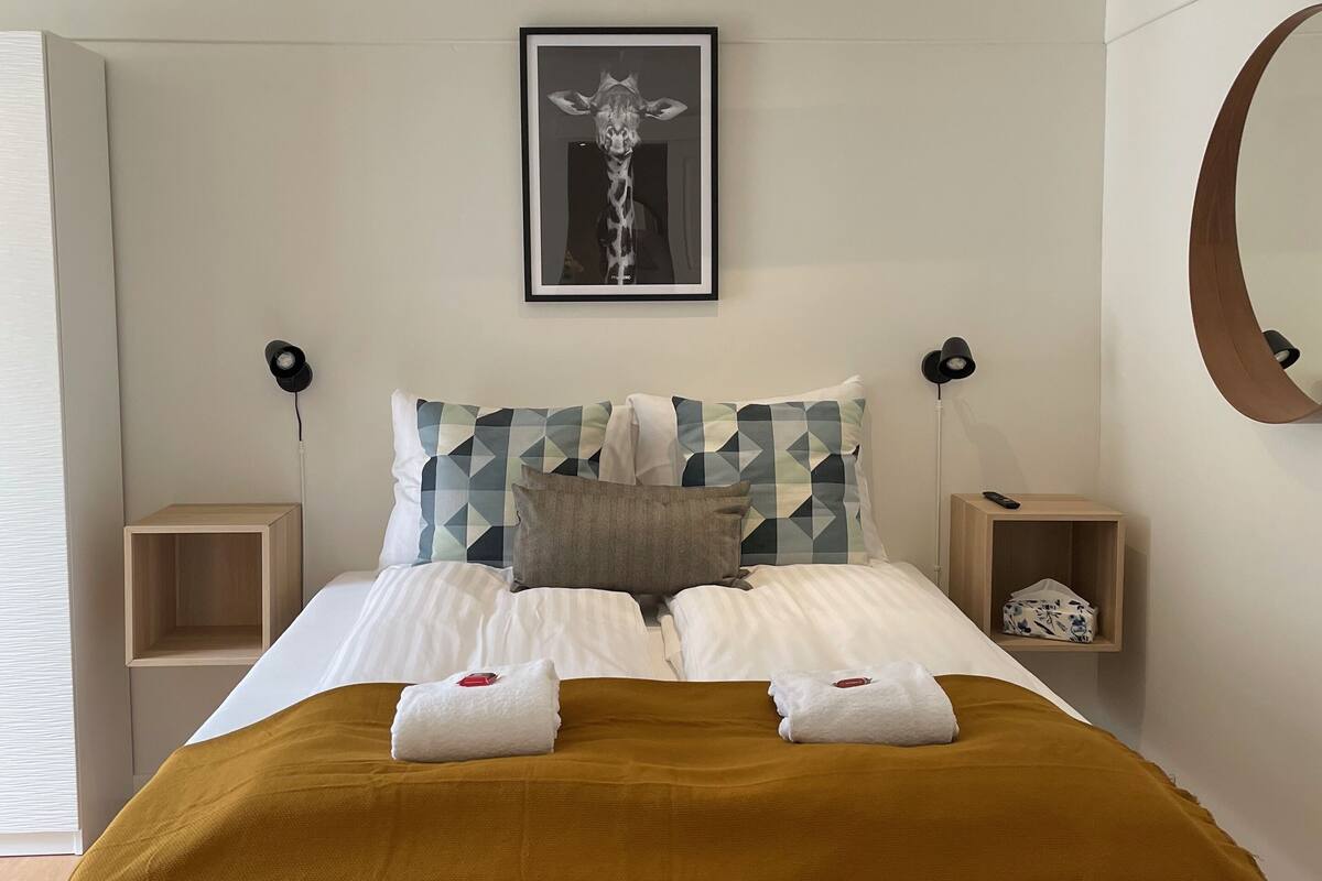 A modern hotel room with a large bed, two bedside tables, a framed picture on the wall, and a round mirror.