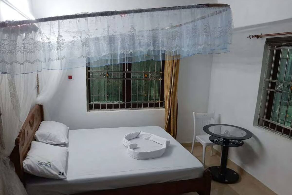 A single bed with a white mattress and pillows, a small round table with a black base, and a window with metal bars and a curtain.