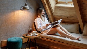 Una mujer sentada en un banco de madera en un espacio tipo loft con techo inclinado, una pequeña mesa con una taza y un platillo, y un otomano verde.