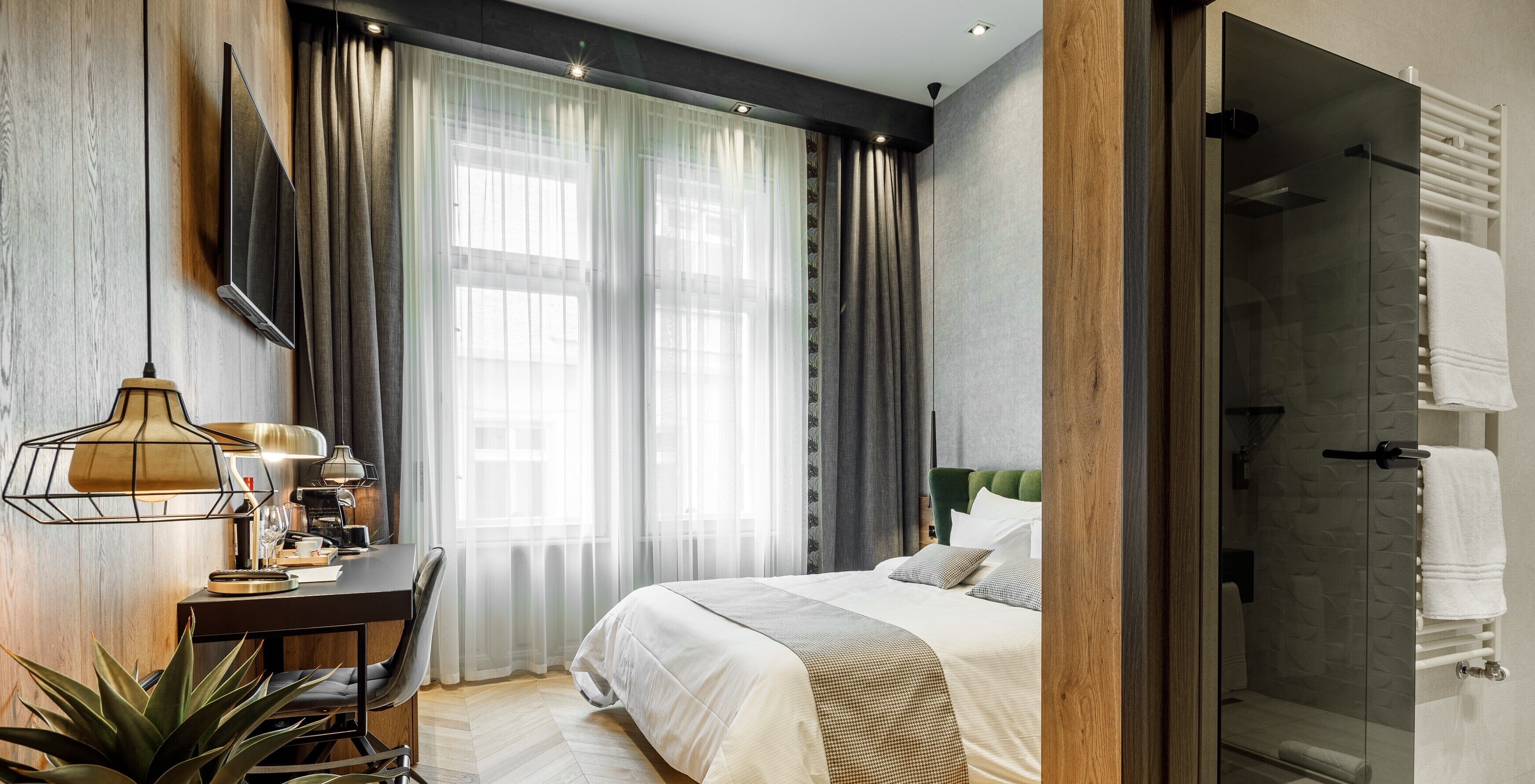 A modern hotel room with a bed, a desk with a lamp, a potted plant, and a shower area.
