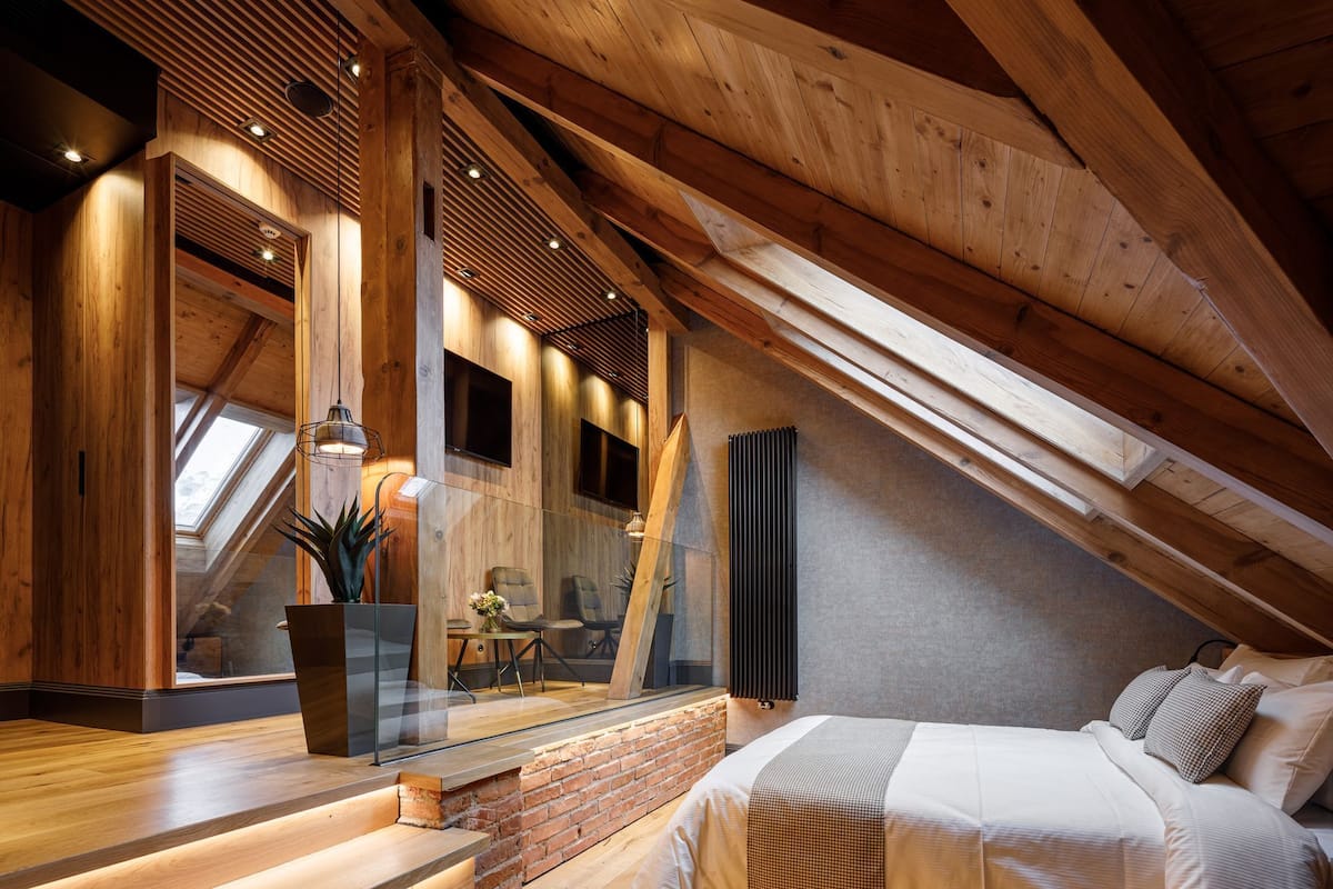 A modern loft bedroom with a sloped ceiling, wooden beams, a brick accent wall, a large bed, and a glass partition with a view of the outside.