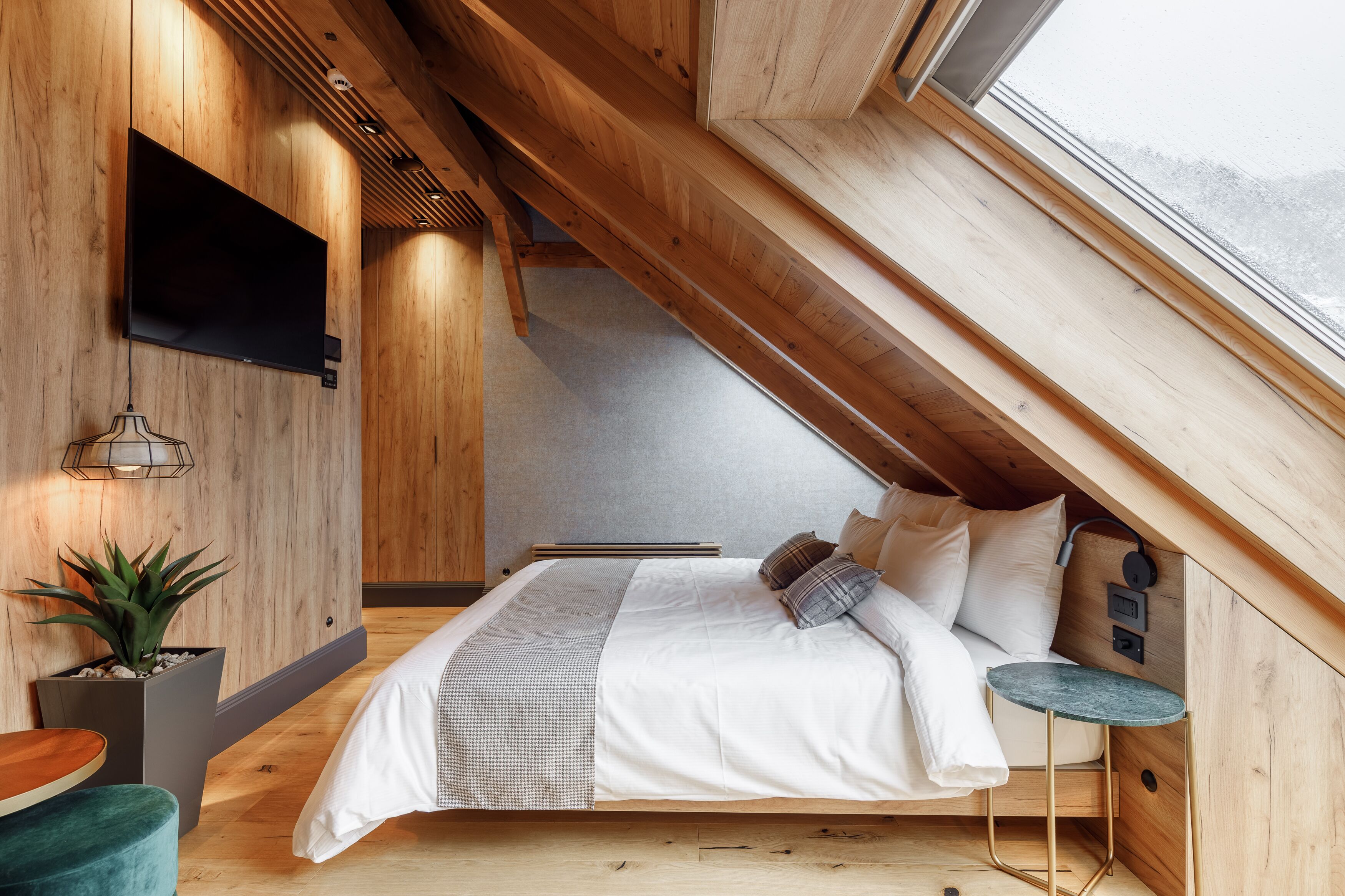 A modern bedroom with a sloped ceiling, a bed with white bedding, a small round table, and a mounted TV.