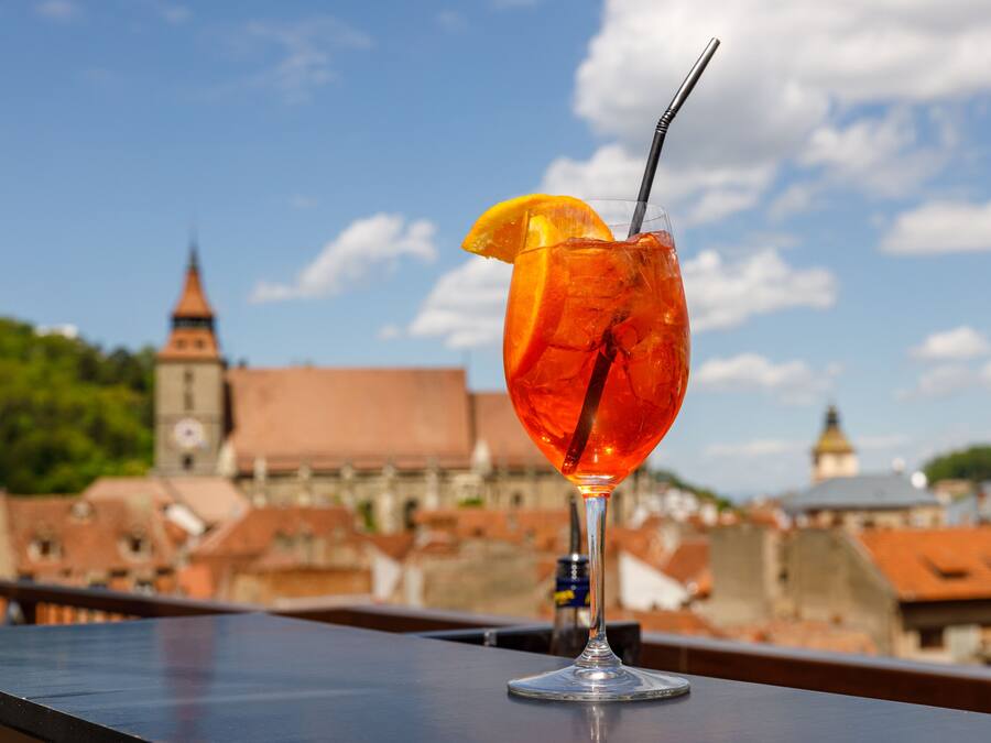 Un vaso de cóctel con una rodaja de naranja en un balcón con vista a un paisaje urbano con edificios históricos.