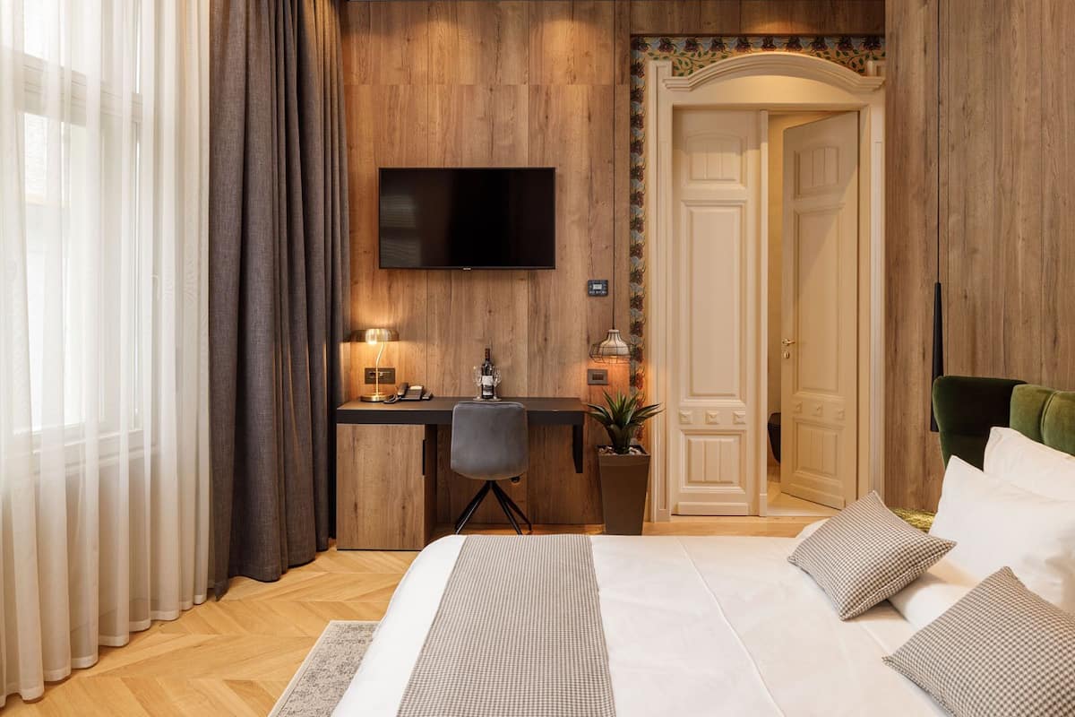 A hotel room with a wooden wall, a flat-screen TV mounted above a desk, a bed with a patterned bedspread, and a green upholstered chair.