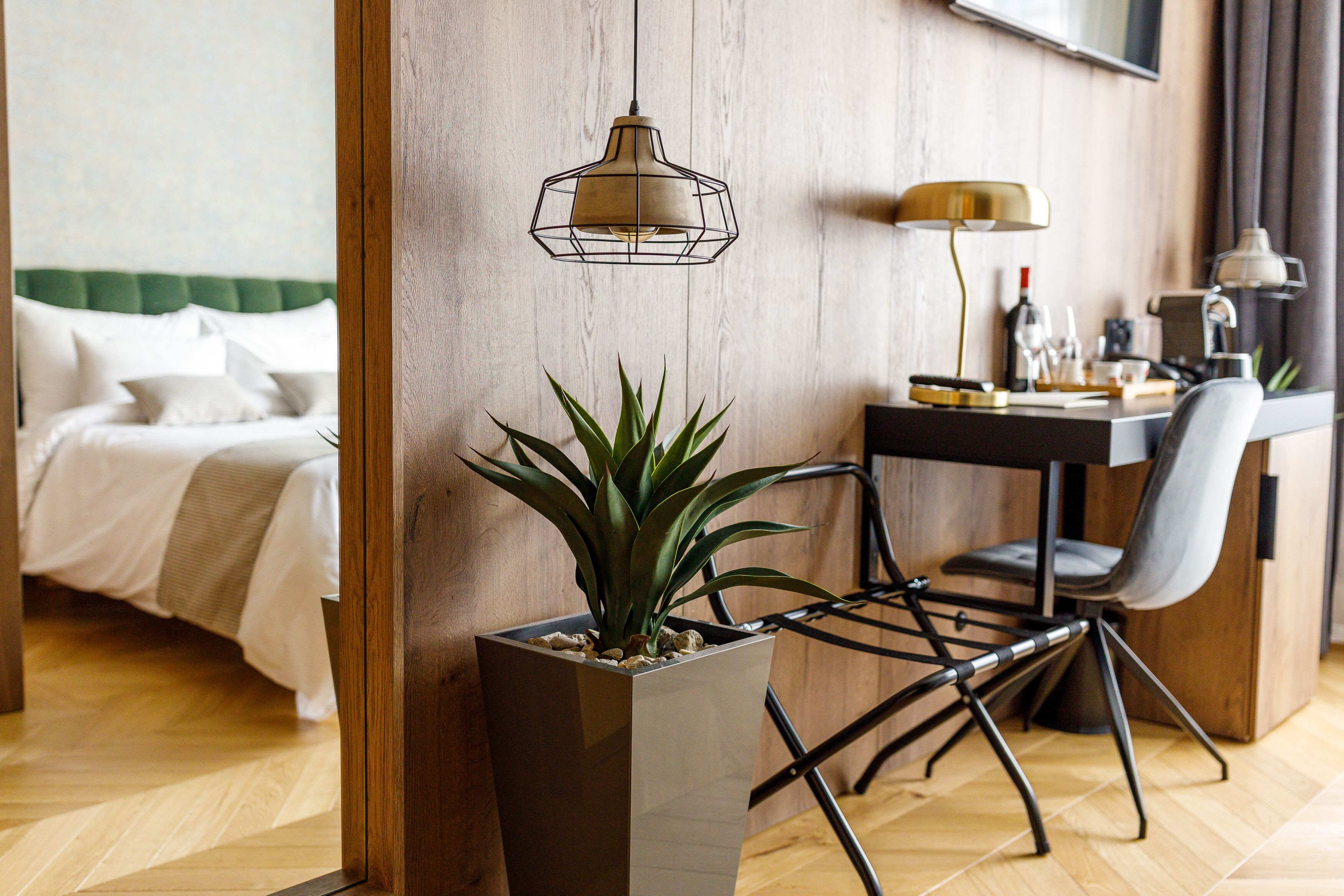 A modern hotel room with a bed, a desk with a chair, a potted plant, and a hanging light fixture.