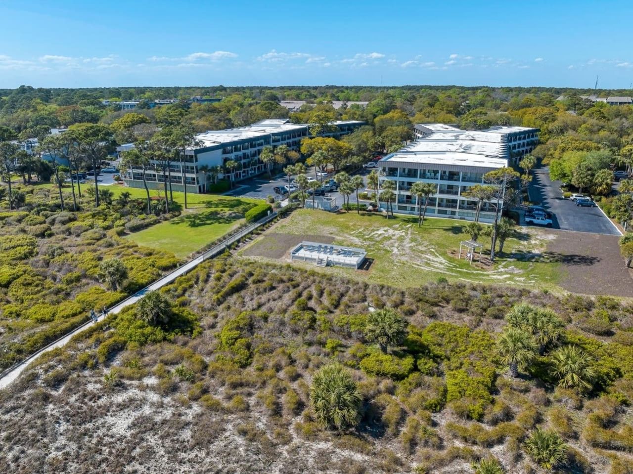 An aerial view of a modern building complex with multiple structures, surrounded by dense vegetation and a clear sky.