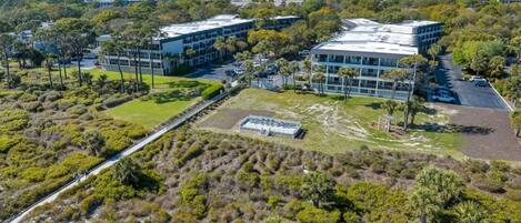 An aerial view of a modern building complex with multiple structures, surrounded by dense vegetation and a clear sky.