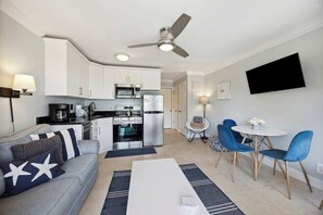 A compact living space with a kitchenette, a dining area with blue chairs, a white sofa with star-patterned pillows, and a ceiling fan.