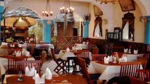 A restaurant with wooden tables and chairs, white tablecloths, and hanging light fixtures.