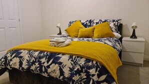Ein ordentlich bezogenes Bett mit einer blumigen Tagesdecke, zwei gelben Kissen und einem gefalteten Handtuch. Auf den beiden Nachttischen stehen Lampen.