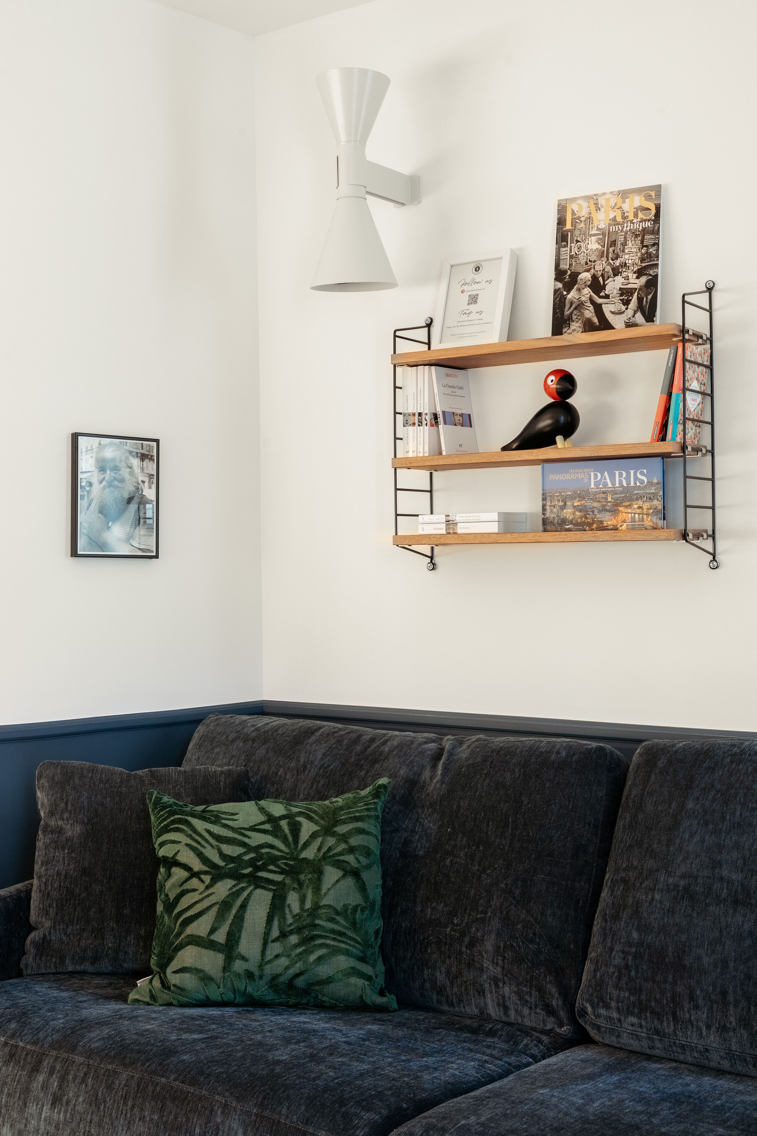 Una sala de estar moderna con un sofá gris oscuro, un cojín estampado en verde, una lámpara colgante blanca y una estantería de pared con libros y una figurita de pájaro.