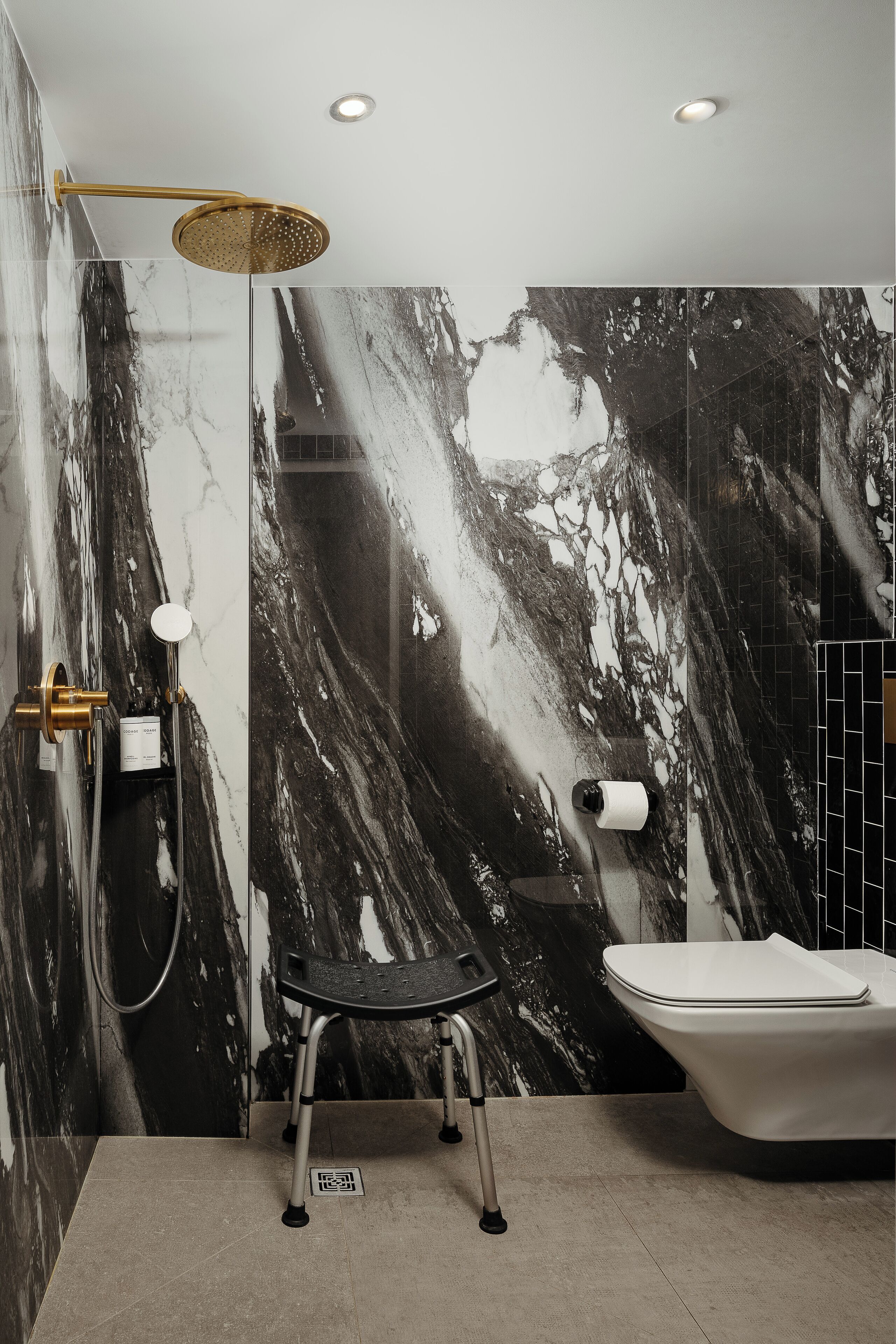 Ein modernes Badezimmer mit Marmorwand, goldfarbener Duschbrause, schwarzem Stuhl und weißer Badewanne.